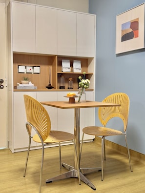 A dining area with a wooden table and two wicker chairs, a cabinet with coffee bags, and a framed abstract painting on the wall. - Runway 1331 - Enlight Youth Hostel (Kowloon)