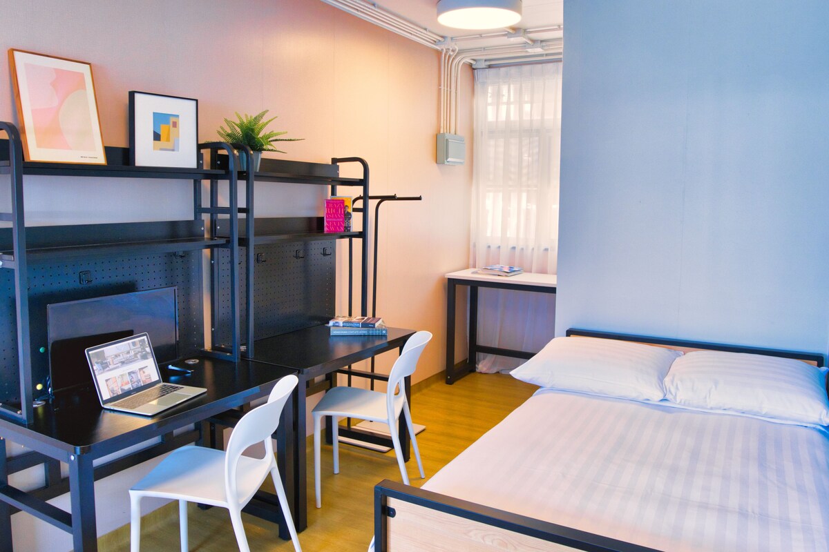 Una habitación compacta con cama, escritorio con laptop, dos sillas, un estante pequeño con libros y una ventana con cortinas.