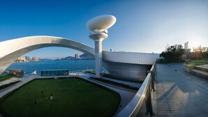 A modern architectural structure with a large arched design and a central tower, overlooking a body of water and a cityscape. - Runway 1331 - Enlight Youth Hostel (Kowloon)