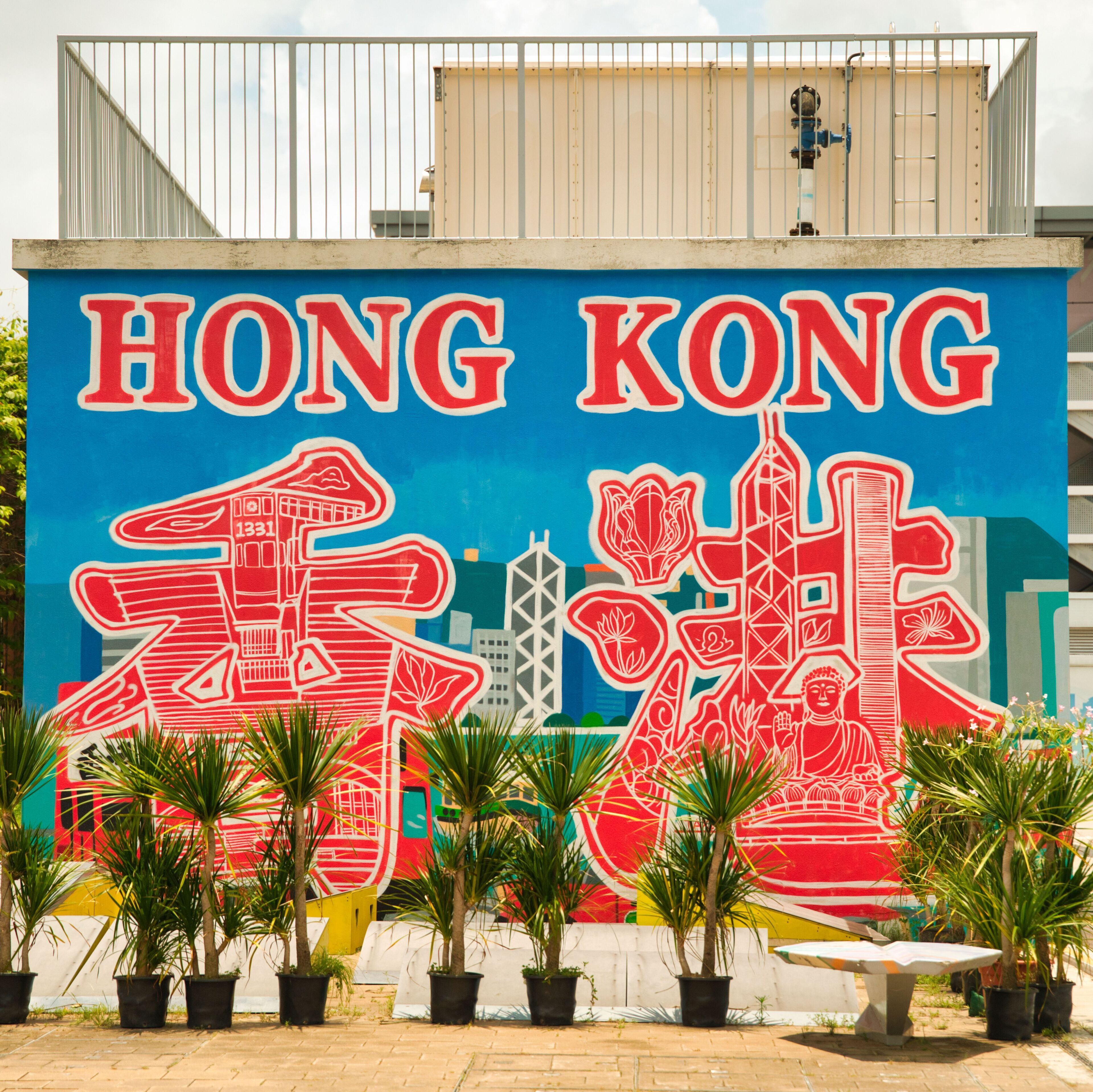 Une fresque murale représentant le mot « HONG KONG » et des bâtiments stylisés, le tout entouré de plantes en pot.
