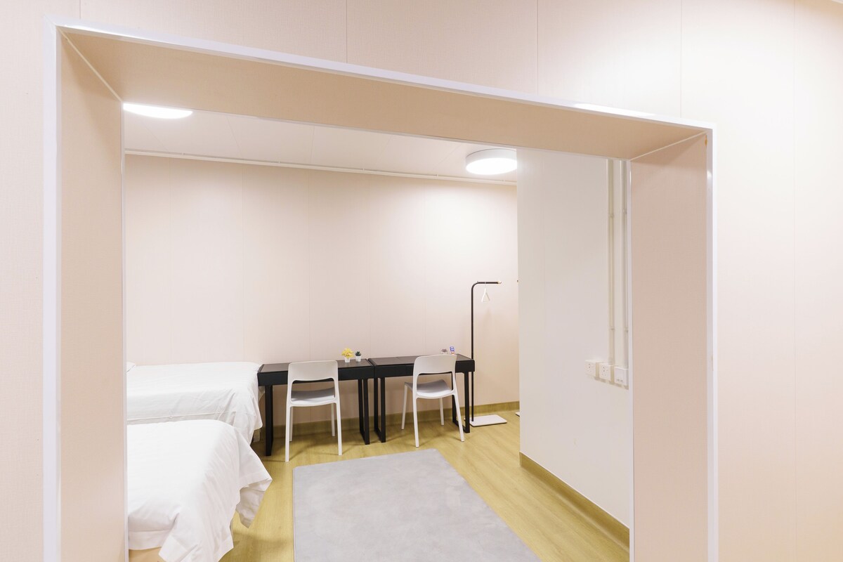 Quarto com cama, escrivaninha com duas cadeiras, abajur e um tapete pequeno.