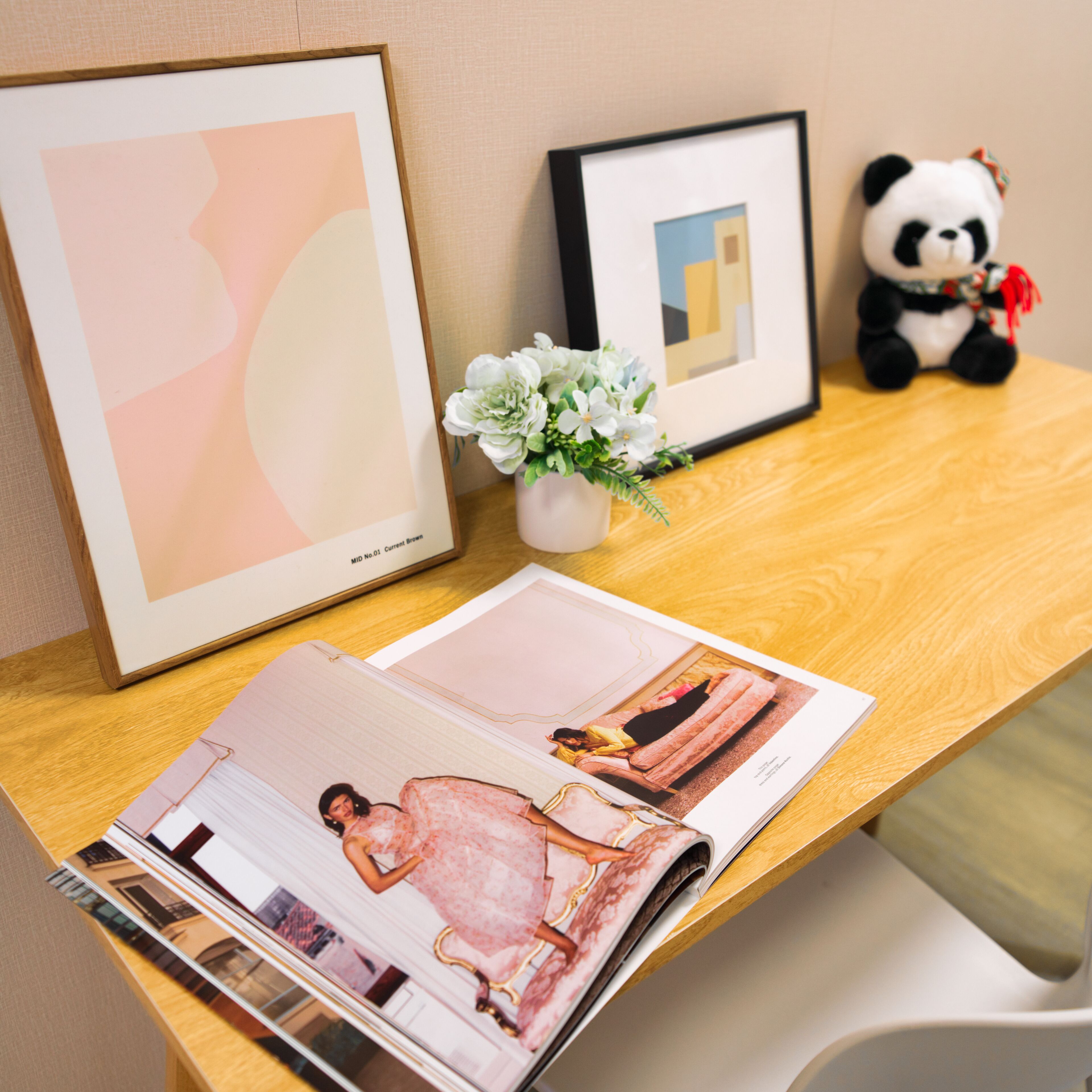 A wooden table with a magazine, a framed abstract painting, a framed geometric painting, a small vase with white flowers, and a panda plush toy.