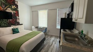 Ein kleines Hotelzimmer mit Bett, einer kleinen Küchenzeile, einem Fernseher und einem Fenster mit Jalousien.