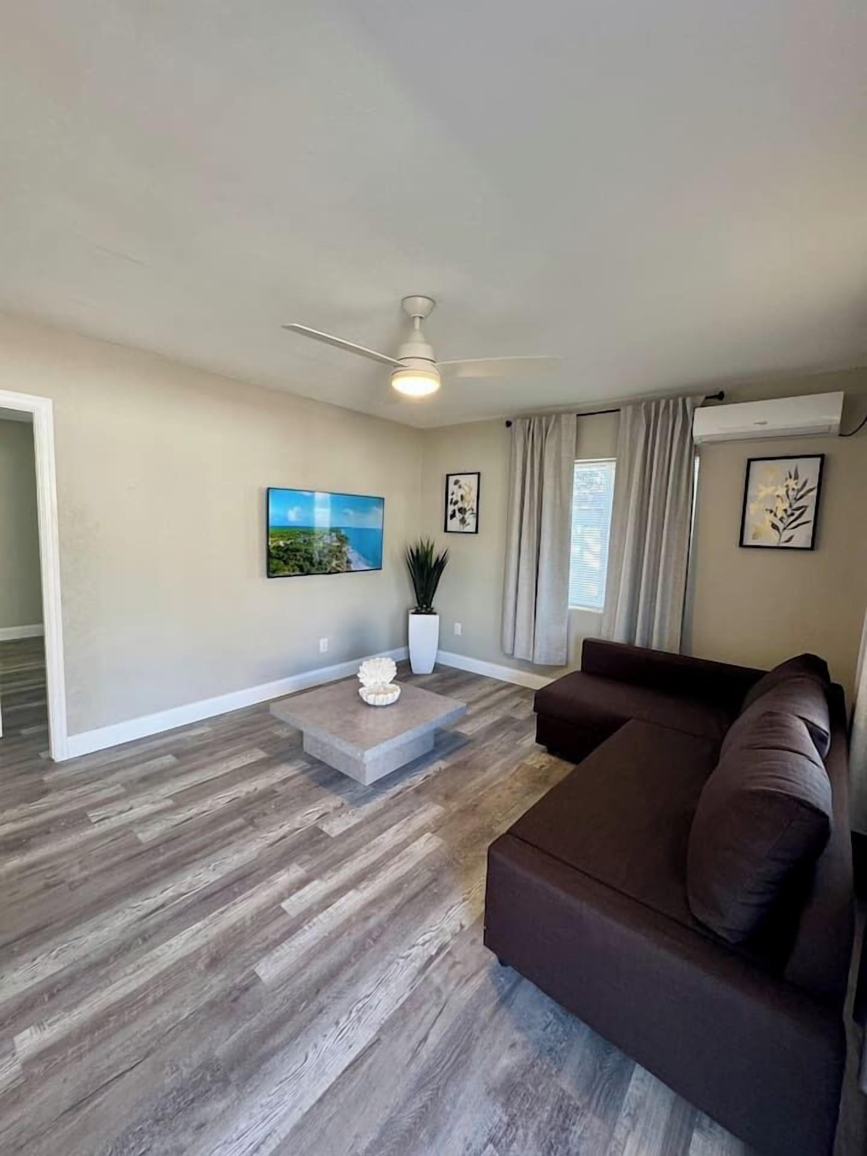 A living room with a brown sofa, a coffee table, a ceiling fan, and a large TV on the wall.