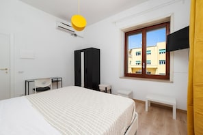 A hotel room with a bed, a desk, a chair, a wardrobe, a window with a view, and an air conditioning unit.
