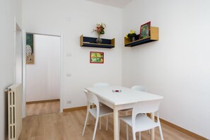 A room with a white table and chairs, a radiator, and wall-mounted shelves with artwork and flowers.