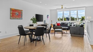 A modern living room with a dining area, large windows, and a flat-screen TV on the wall.