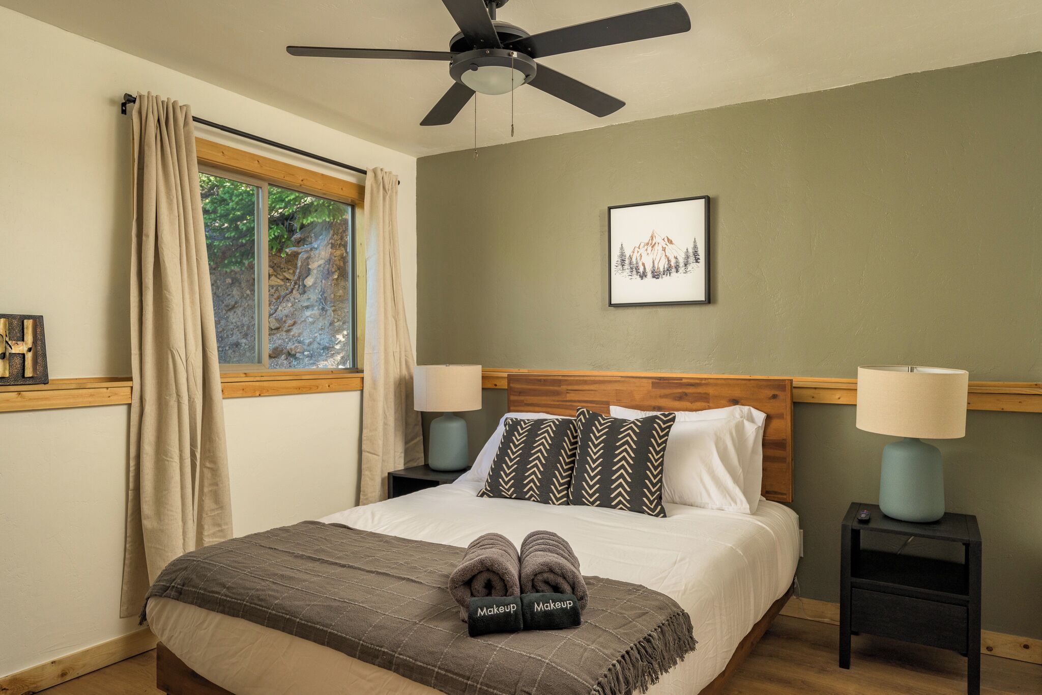 Un dormitorio con cama, dos mesitas de noche con lámparas, ventilador de techo, ventana con cortinas e imagen enmarcada en la pared.