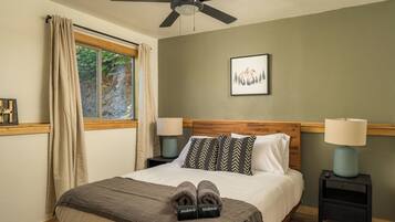 Un dormitorio con cama, dos mesitas de noche con lámparas, ventilador de techo, ventana con cortinas e imagen enmarcada en la pared.
