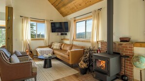 Ein gemütliches Wohnzimmer mit Ledersofa, Fernseher, Kamin und einem großen Fenster mit Blick ins Freie.