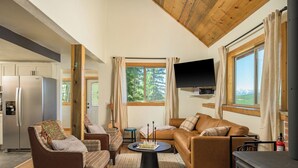 Ein gemütliches Wohnzimmer mit Sofa, Sesseln, einem Couchtisch und Fernseher. Es gibt ein Fenster mit Vorhängen, einen Kamin und eine Küchenzeile mit Kühlschrank.