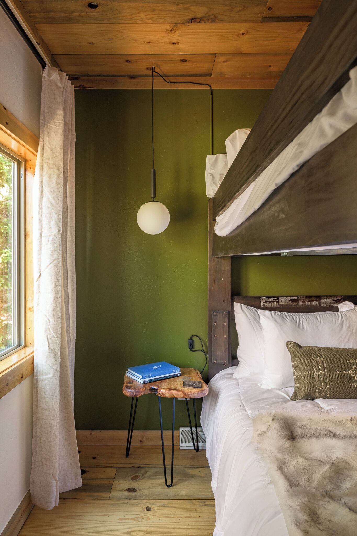 A bunk bed with a wooden headboard, a small table with a book, and a hanging light fixture in a room with green walls and wooden floors.