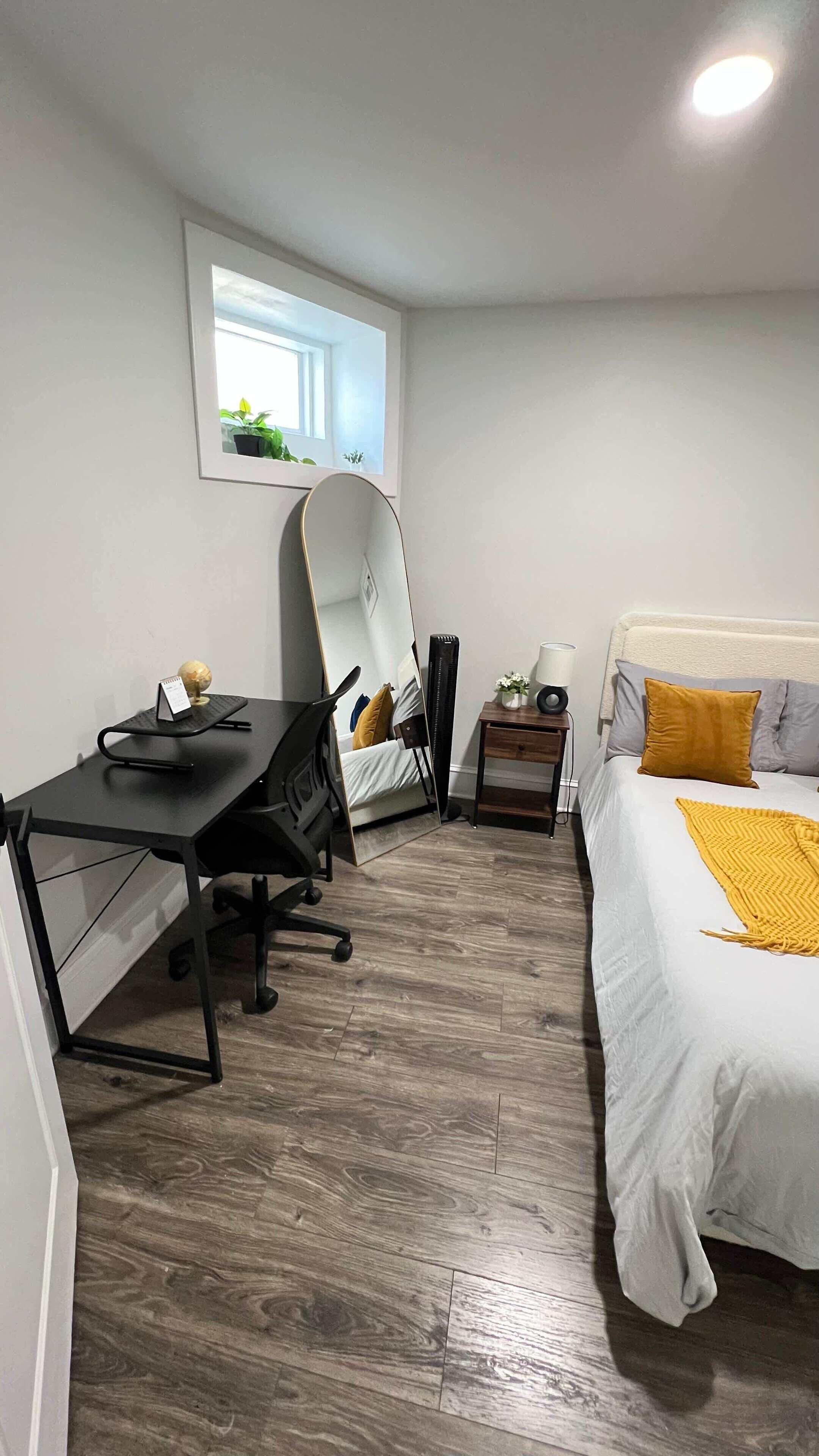 A compact bedroom with a bed, a desk, a chair, a mirror, a bedside table, and a window.