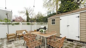 A patio with a wooden table and wicker chairs, a barbecue grill, and a shed in the background.