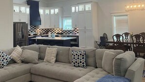 A modern living room with a sectional sofa, a dining area with a wooden table and chairs, and a kitchen with white cabinets and a black and white backsplash. - AG Ranch in Hempstead (Hempstead)