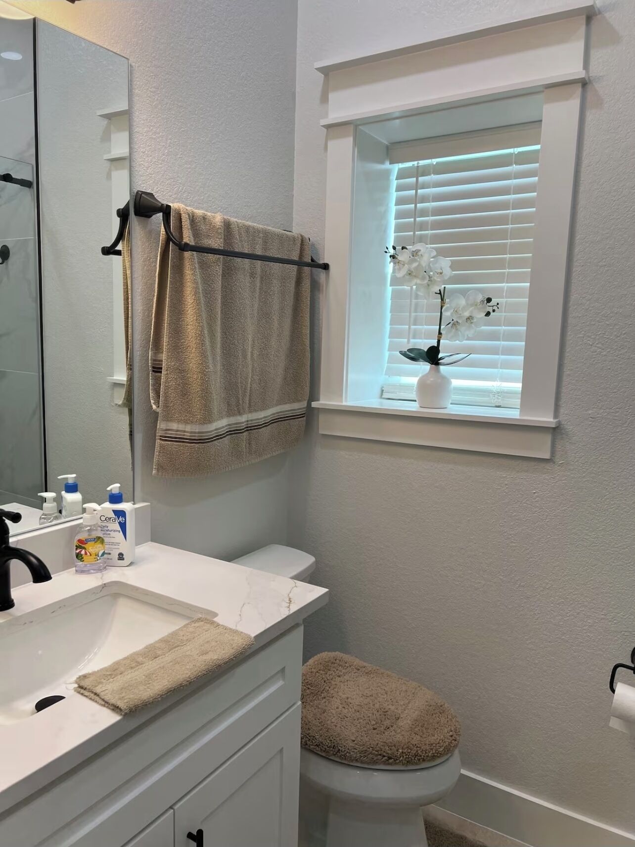 A bathroom with a white sink, a toilet, a towel rack with a towel, a window with blinds, and a vase with flowers.