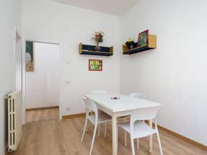 Une pièce avec une table blanche, deux chaises blanches, un radiateur et des étagères murales ornées d’objets décoratifs.