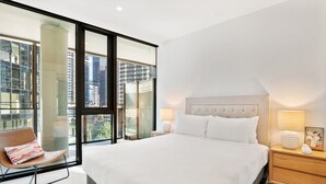 A modern hotel room with a large bed, a chair, a nightstand, and a view of the city through floor-to-ceiling windows. - Spacious Modern Apartment With Pool & Gym Access (Melbourne)