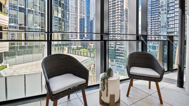 A modern balcony with two wicker chairs, a small white side table with a plant, and a city view through large windows. - Spacious Modern Apartment With Pool & Gym Access (Melbourne)