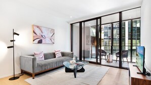 A modern living room with a grey sofa, a glass coffee table, and a large window with a city view. - Spacious Modern Apartment With Pool & Gym Access (Melbourne)