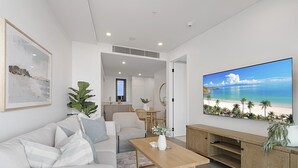 Een moderne woonkamer met een grote televisie waarop een strandscène te zien is, een bank, een salontafel, en een eethoek met stoelen en een tafel.