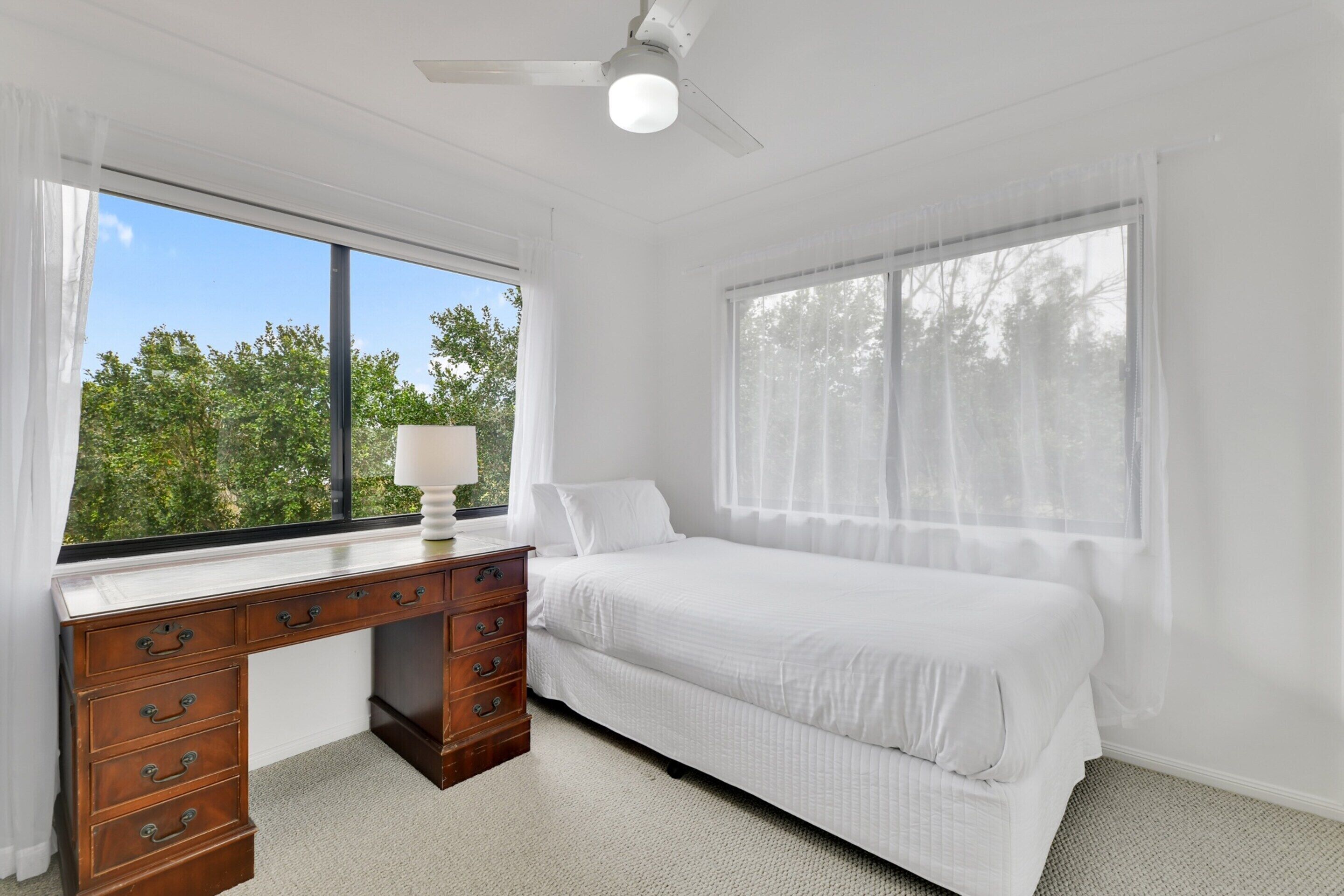 A bedroom with a wooden desk, a bed, and a window with a view of trees.
