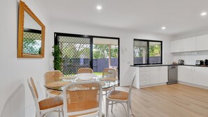 A modern kitchen with white cabinets and a dishwasher, a dining area with a glass table and wicker chairs, and a large window with a view of a backyard. - Homely 3-bed Townhouse With Pool & Gym Access (Brisbane)