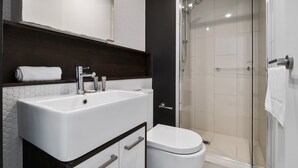 Un baño moderno con lavamanos, espejo y retrete blancos, y una zona de ducha con cerramiento de vidrio.