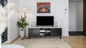 Una sala de estar moderna con un televisor de pantalla plana, una consola con libros, una lámpara de pie y un cuadro decorativo.