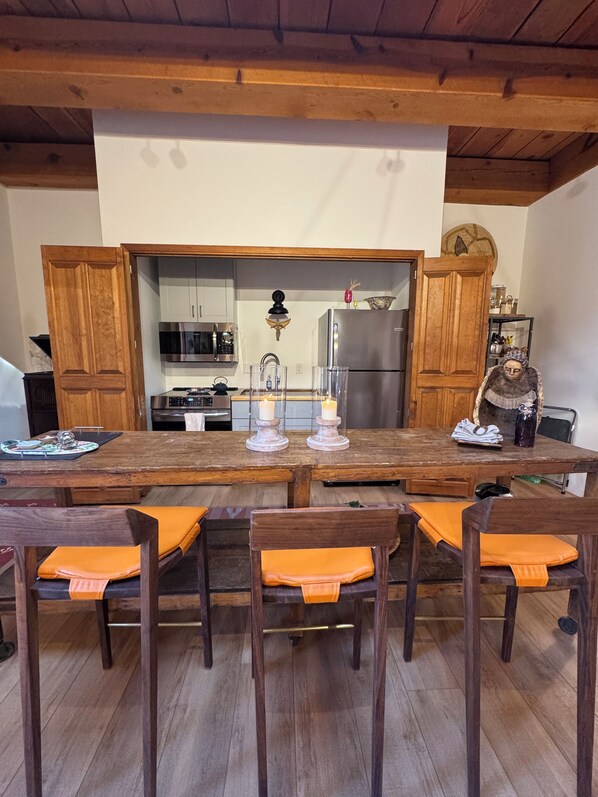 A wooden dining table with orange cushions, a kitchen with wooden cabinets, a refrigerator, and a microwave.