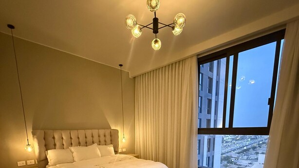 A modern bedroom with a large bed, bedside tables, a chandelier, and a view of the city through large windows.
