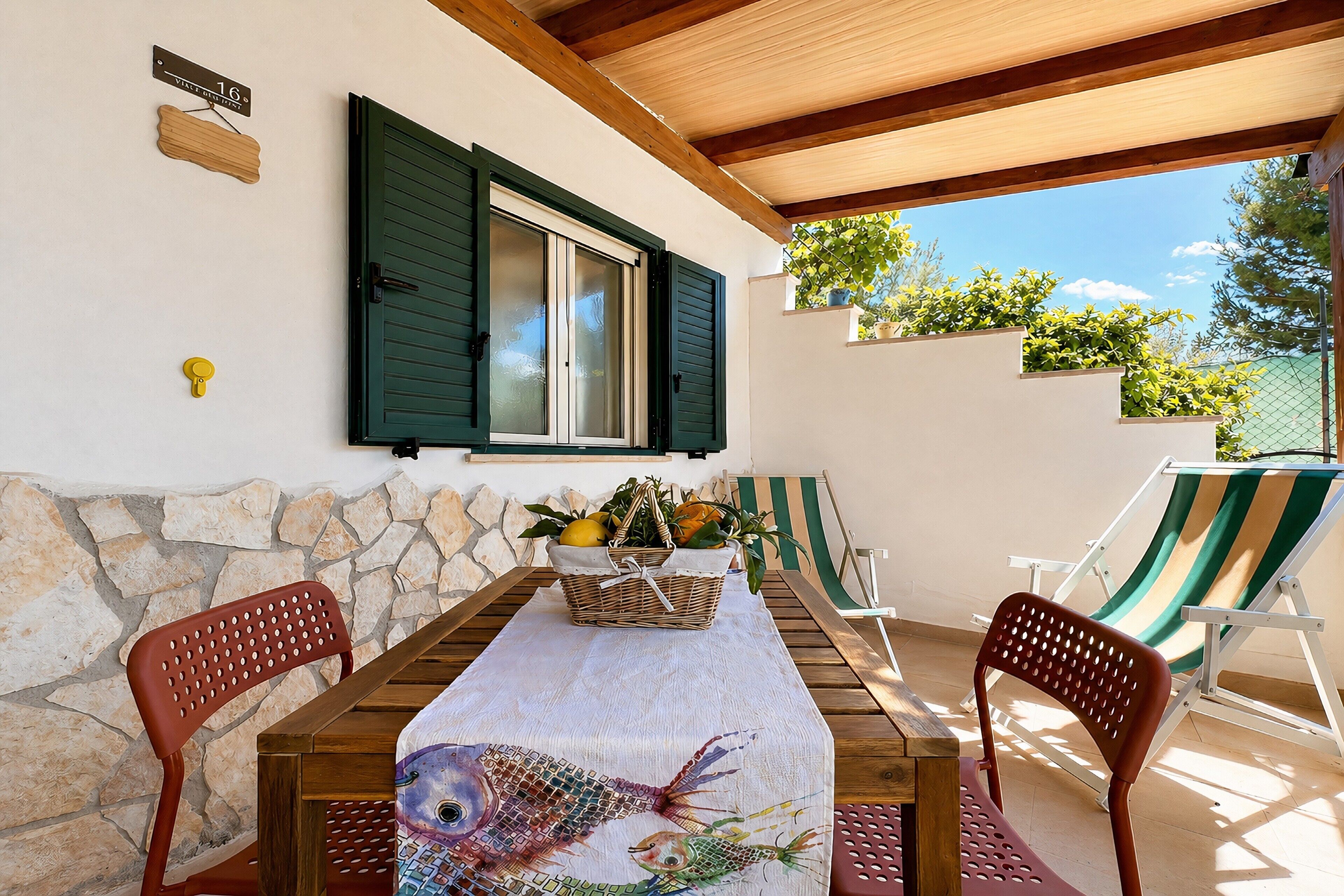 A covered patio with a table, chairs, a wicker basket with fruits, a fish-patterned tablecloth, and a stone wall.