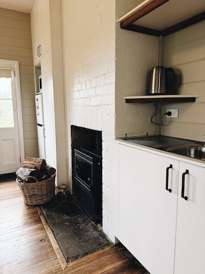 Una cocina con pared de ladrillo blanco, estufa negra, suelo de madera y un hervidor de metal sobre una estantería.