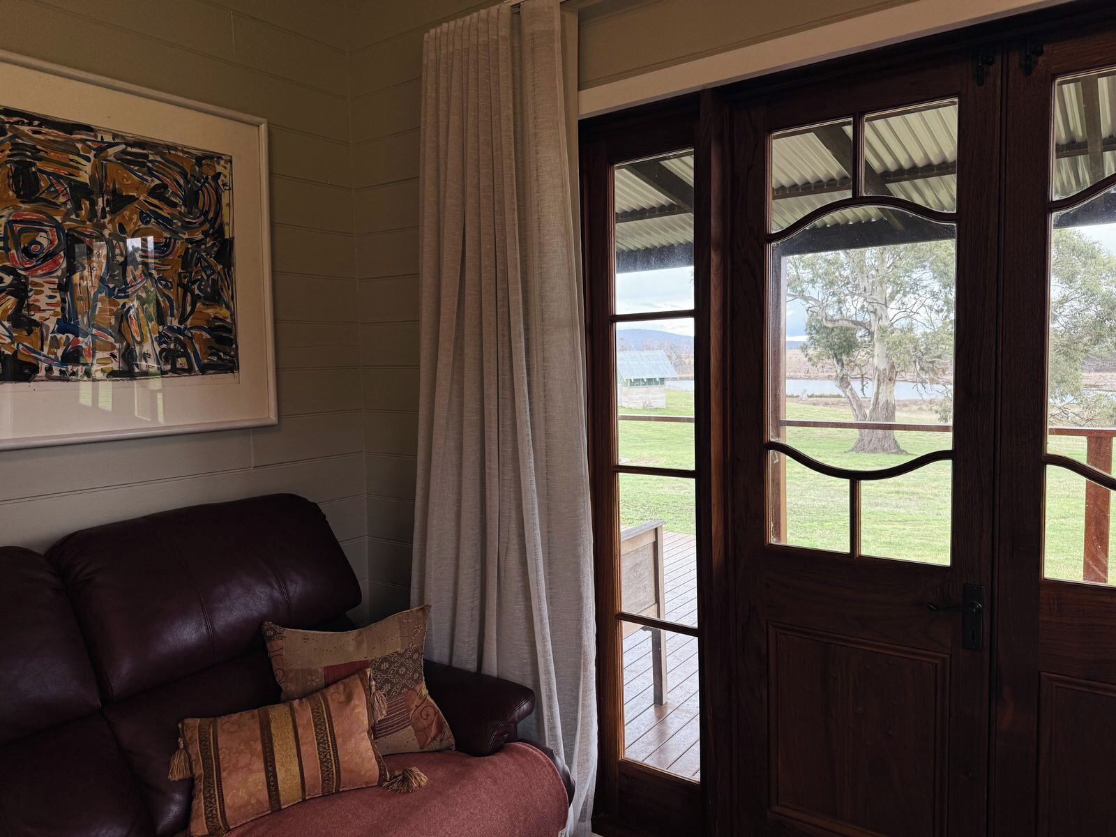 A room with a leather sofa, a framed abstract painting, and a view of a grassy area through a wooden door with glass panels.