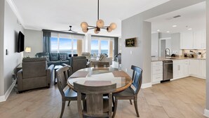 A spacious living room with a round wooden table and chairs, a modern chandelier, and a view of the ocean through large windows. - Beach Club B1805 Pools-54-ft balcony K/K/K/K/Bonus room with Q & Tw-XL Sleeps 11 (Gulf Shores)