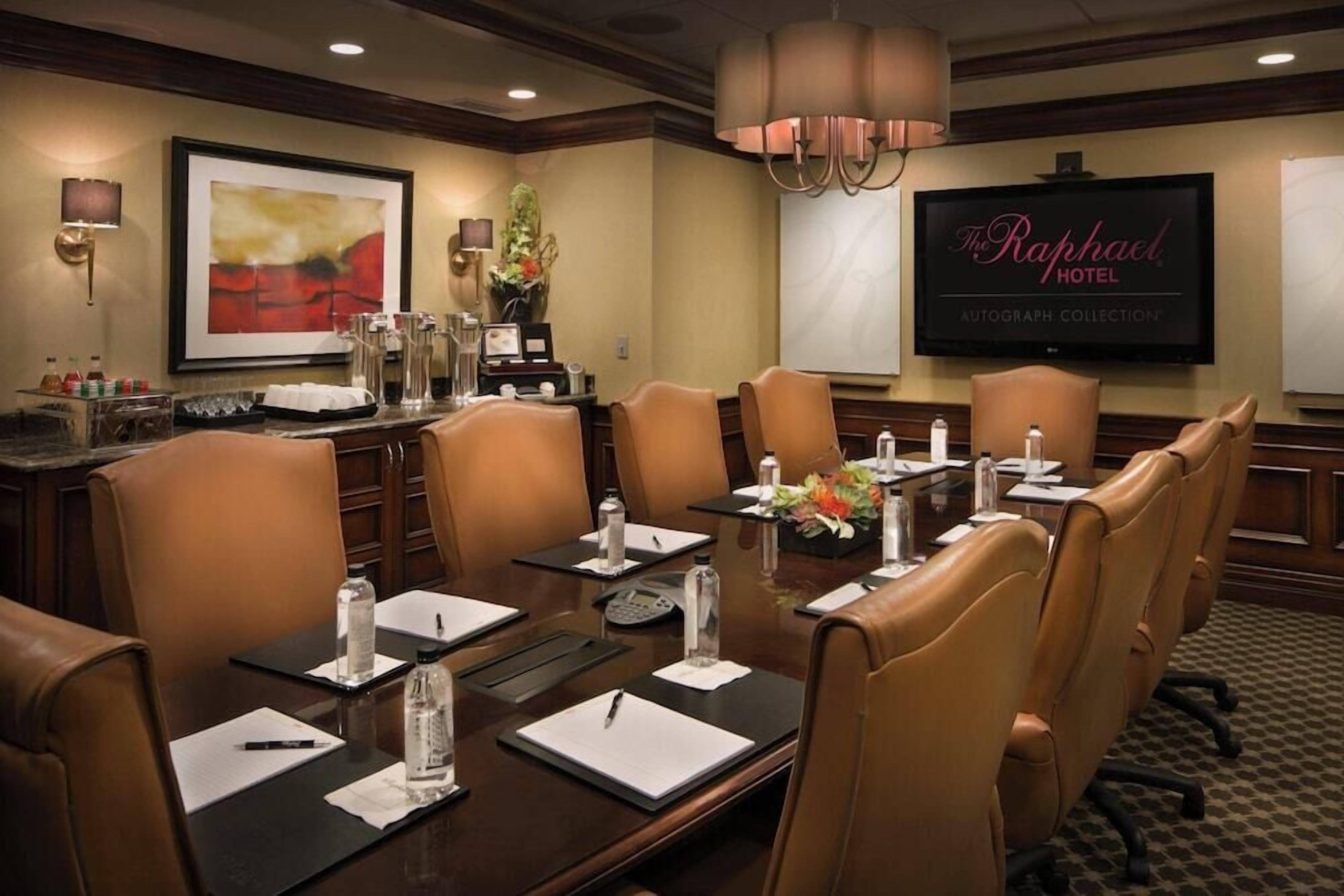 Un salón de conferencias con una gran mesa, sillas de cuero, botellas de agua, blocs de notas, bolígrafos y una televisión mostrando 'The Raphael Hotel Autograph Collection'.
