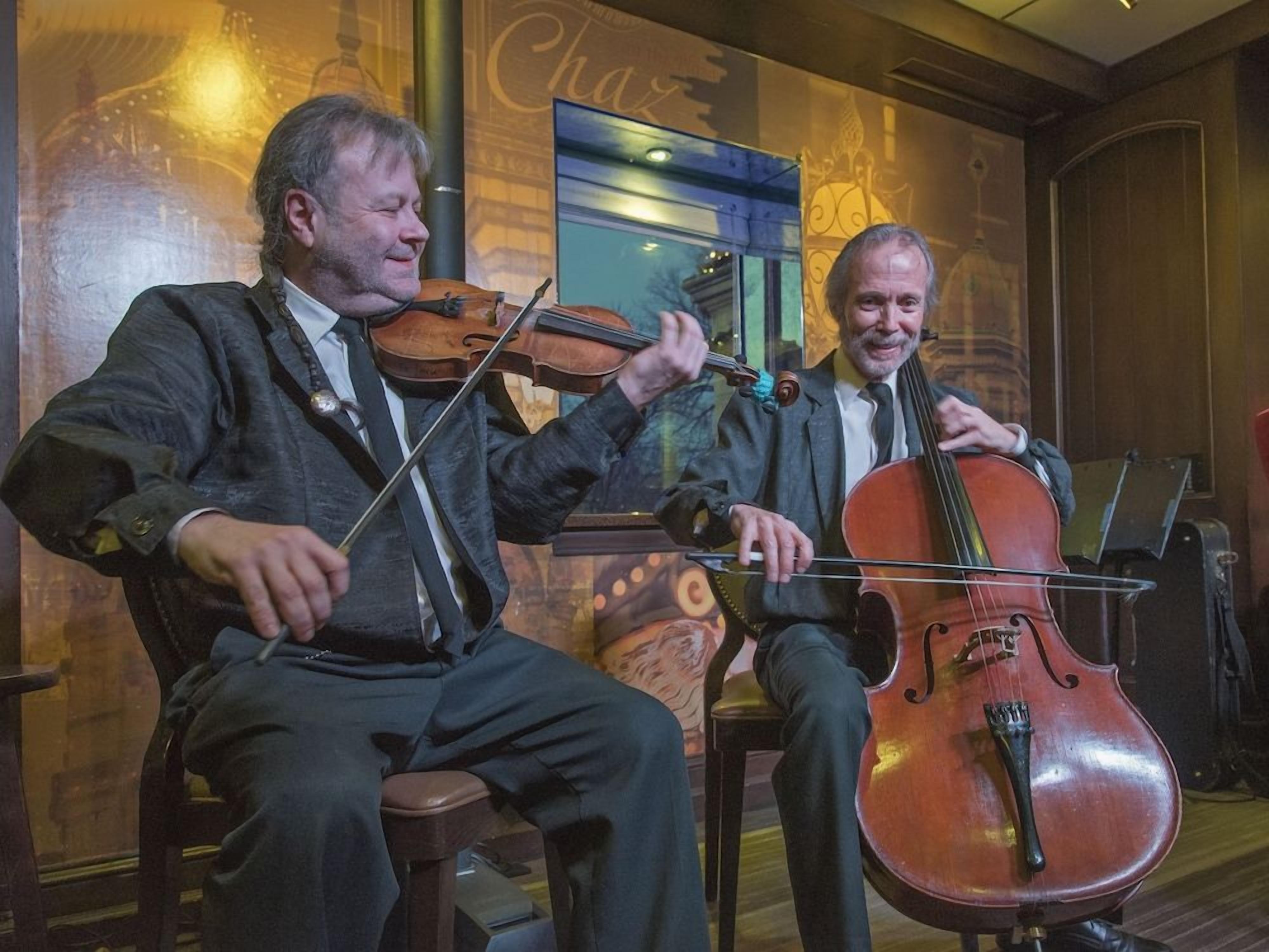 Dos músicos sentados, uno tocando el violín y el otro el violonchelo, en una habitación con paredes de madera y una ventana con reflejo.