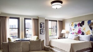 A hotel room with a large bed, a sofa, a desk, and a view of the city through large windows.