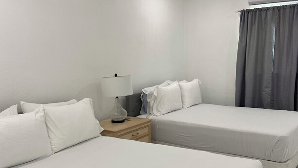 Habitación de hotel con dos camas, una mesita de noche con lámpara, aire acondicionado de pared y ventana con cortinas.