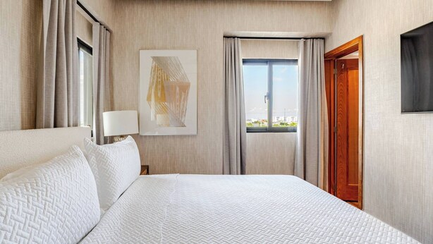 A hotel room with a large bed, a window with curtains, a painting on the wall, and a wooden door.
