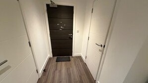 Un pasillo estrecho con una puerta de madera oscura al final, paredes blancas y suelo de madera.