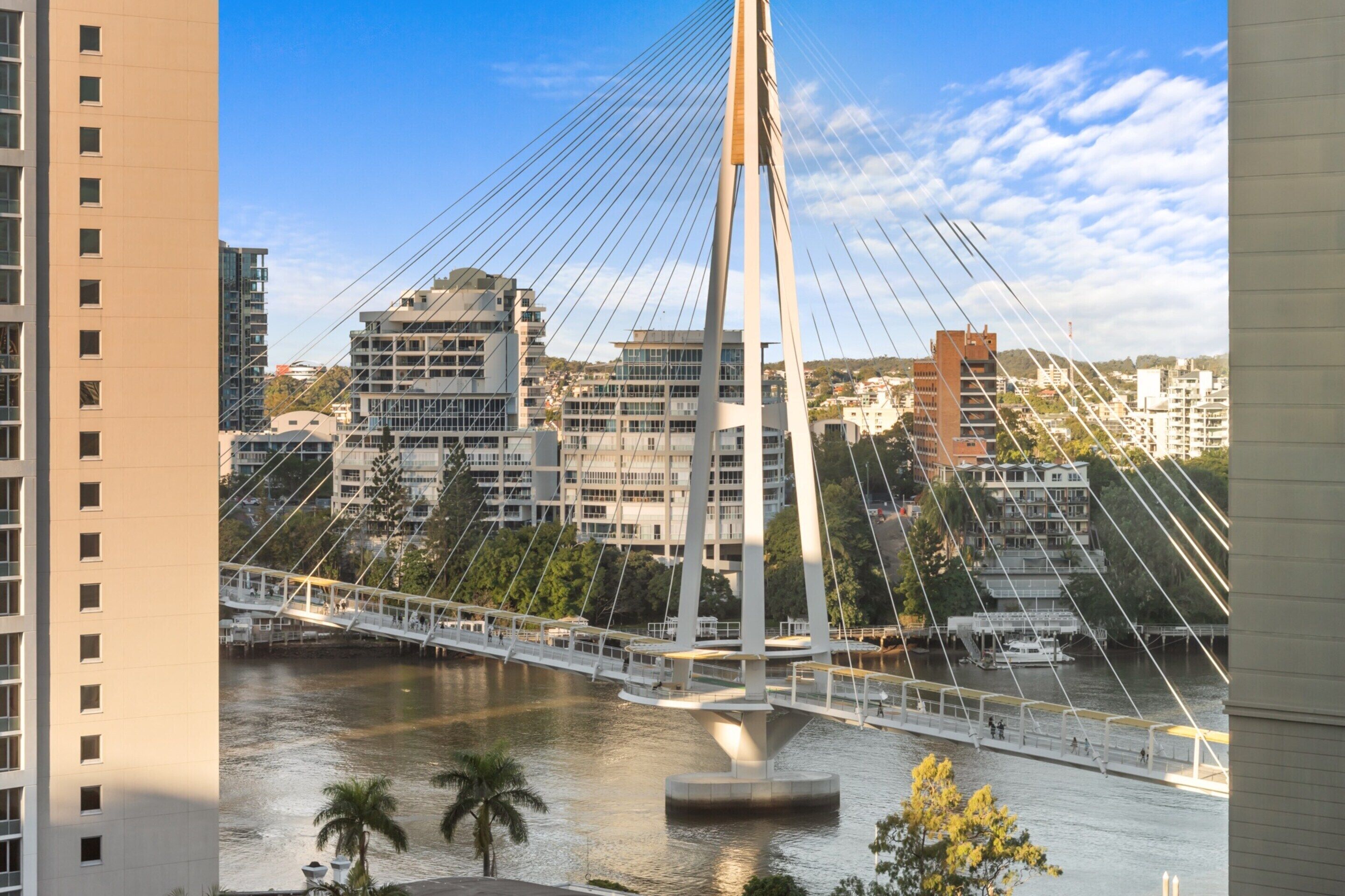 A modern cable-stayed bridge with a pedestrian walkway over a river, surrounded by urban buildings.