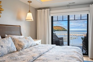 A bedroom with a large bed, a nightstand, a hanging lamp, and a view of a body of water and a distant island.