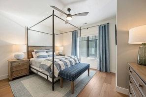 A four-poster bed with a ceiling fan, a bedside table with a lamp, a bench, a window with curtains, and a dresser.