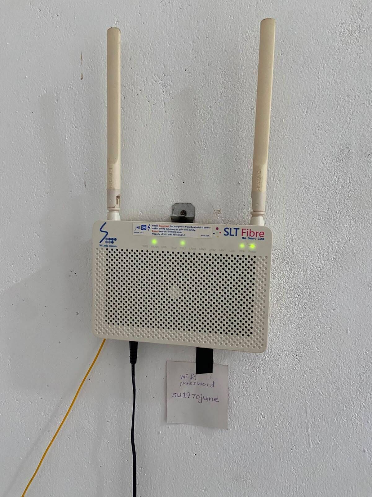 A white SLT Fibre router with two antennas, a power cable, and a label with a Wi-Fi password.