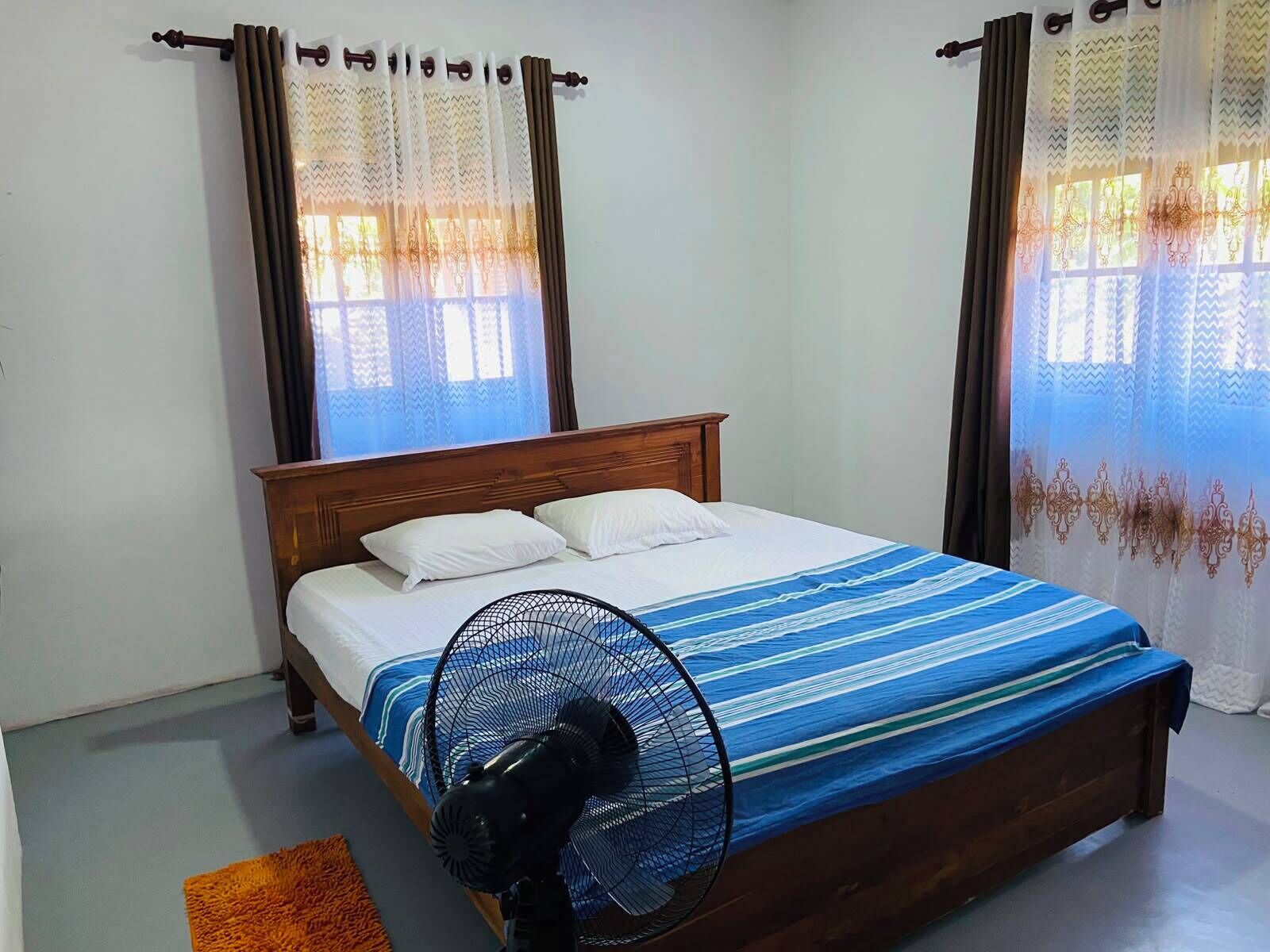 A bedroom with a wooden bed, white pillows, a blue-striped bedspread, a standing fan, and curtains with a zigzag pattern.