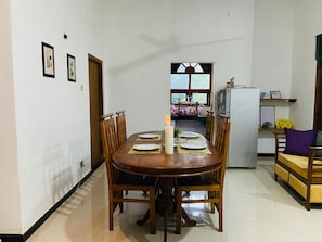 A dining area with a wooden table set for four, a candle, and chairs. A kitchen is visible in the background with a refrigerator and a window.