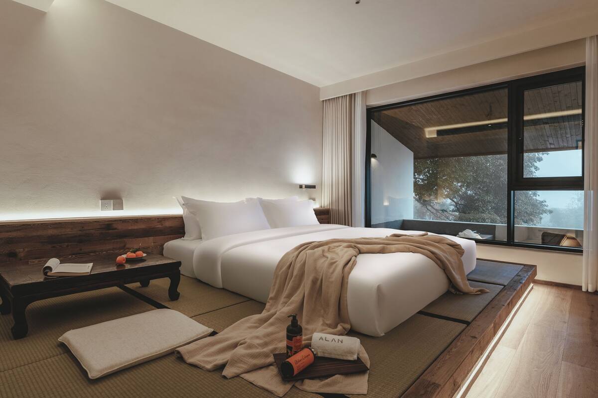 A modern hotel room with a large bed, a wooden bench, a small table, and a view of the outdoors through a large window.