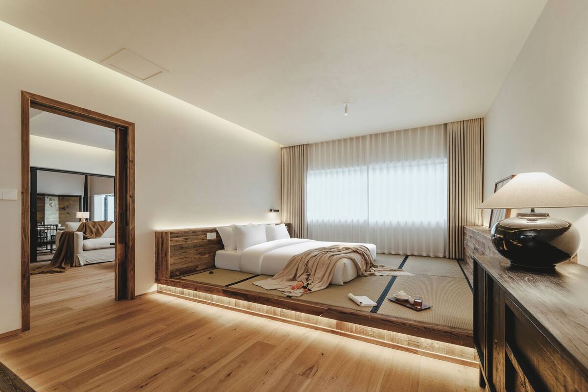 A modern hotel room with a wooden floor, a bed with a headboard, a bedside table with a lamp, and a large mirror.