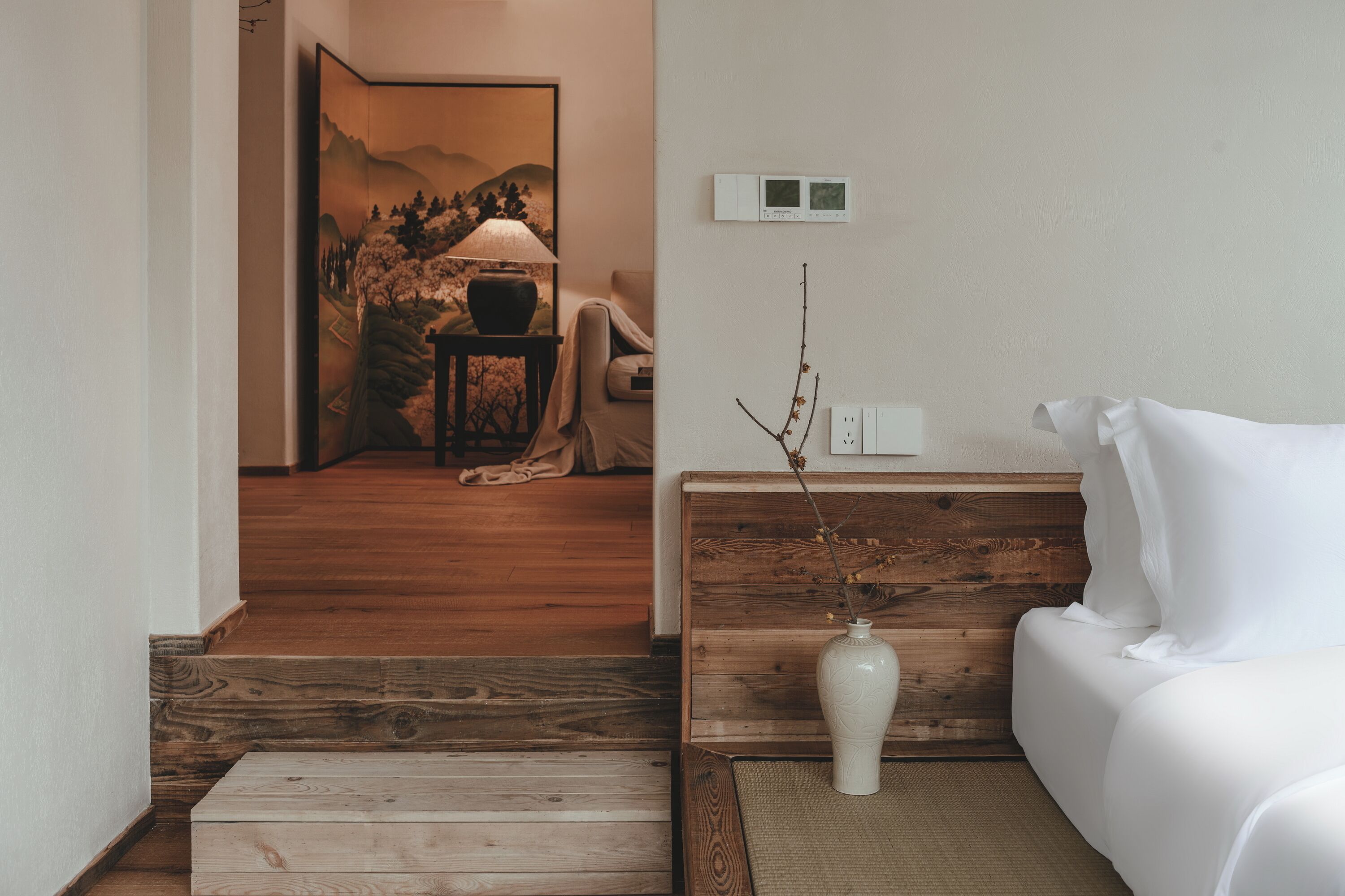 A room with a wooden floor, a white sofa, a wooden headboard, a white vase with branches, a lamp, and a painting on the wall.